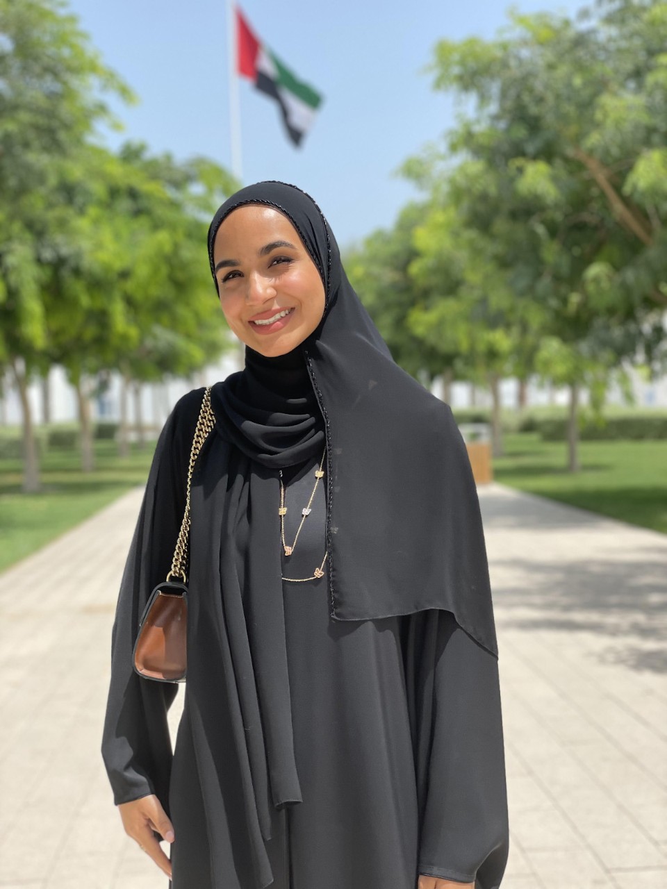 Maryam Alshehhi, standing outdoors on a tree-lined athway, with the UAE flag in the background.
